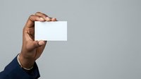 Male hand with dark skin tone holding blank white business card against neutral gray background for corporate branding and marketing mockups.