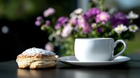 Fresh baked pastry with powdered sugar next to white porcelain coffee cup and saucer on dark surface, purple flowers blooming in soft focus background.
