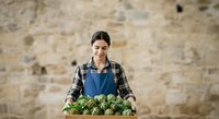 Young woman with dark hair and South Asian ethnicity wearing blue apron and plaid shirt holding crate of fresh broccoli against rustic stone wall. Farmer, gardener, produce seller.
