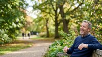 Middle-aged man with light skin tone and gray hair, wearing a navy sweater, laughing while sitting on a bench in a lush green park with tree-lined pathways, suitable for lifestyle, wellness, and outdoor recreation content.