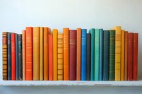 Row of vintage hardcover books arranged by color from red to orange to blue to green on white shelf, creating rainbow gradient effect for library decor.