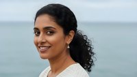 Happy young South Asian woman with curly hair smiling at camera near ocean wearing white top for lifestyle and wellness content.