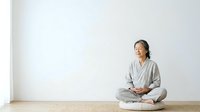 Woman in casual gray clothing sitting cross-legged on wooden floor against white wall, meditating or practicing mindfulness in minimalist interior space.