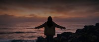 Silhouetted figure with arms outstretched standing on rocky coastline at sunset, experiencing freedom and contemplation by the sea during golden hour.