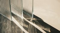 Sunlight streams through vertical blinds casting geometric shadows on a neutral wall and floor, creating an abstract interplay of light and dark stripes for modern interior design projects.