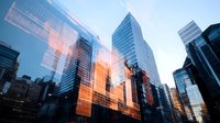 Modern glass skyscrapers reflecting golden sunset light against blue sky, creating dramatic urban architecture with reflective surfaces and geometric patterns.
