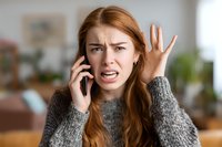 Frustrated young Caucasian woman with red hair talking on smartphone, expressing anger and confusion with hand gesture in modern home interior.