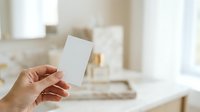Woman's hand holding blank white card in modern kitchen interior with natural light for branding, business card mockup, or promotional material display.