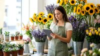 Young female florist in apron using tablet in bright flower shop surrounded by sunflowers and lavender plants for small business management.