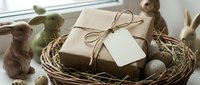 Eco-friendly gift box with blank tag in wicker basket surrounded by wooden Easter bunnies and speckled eggs on windowsill for spring celebration.
