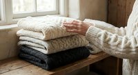 Woman in cream knit sweater organizing stack of cozy winter sweaters on wooden table by window for seasonal wardrobe storage and home organization.