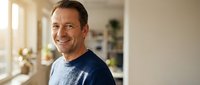 Smiling middle-aged Caucasian man in casual blue sweater standing in bright modern home interior with natural sunlight streaming through windows.