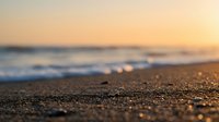 Sandy beach shore with soft waves and golden sunset light creating bokeh effect on wet sand and ocean backdrop.