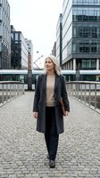 Professional blonde businesswoman walking confidently through modern urban district wearing elegant coat and carrying leather portfolio.