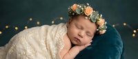 Peaceful newborn baby girl sleeping wrapped in cream lace blanket wearing floral crown headband on teal backdrop with warm bokeh lights for nursery decor.