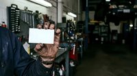 Mechanic with tattooed arms holding blank white sign in industrial garage workshop surrounded by tools and equipment, wearing work clothes and gloves for automotive repair business messaging.