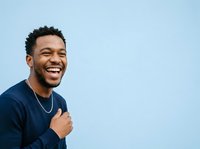 Young African American man with short curly hair wearing navy blue long-sleeve shirt and silver chain necklace, smiling broadly against light blue background. Portrait for lifestyle and advertising use.
