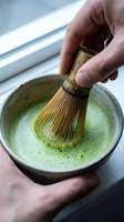 Person whisking matcha tea powder in ceramic bowl with traditional bamboo brush, preparing green tea beverage for culinary and wellness purposes.