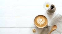 Overhead view of latte with latte art beside fresh daisy flower and wooden spoon on white wooden surface for café, coffee shop, and lifestyle design projects.