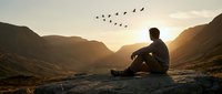Man sitting on mountain rock watching birds fly at golden sunset in peaceful valley landscape for meditation and mindfulness concepts.