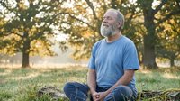 Mature man with gray beard and light skin tone, wearing blue t-shirt and jeans, sitting cross-legged on grass in park during golden hour with trees in background, conveying relaxation and mindfulness.
