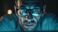 Caucasian male programmer with glasses illuminated by blue computer screen light at night, dramatic portrait showing focused concentration and digital lifestyle.
