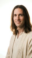 Confident Caucasian man with long brown hair wearing traditional embroidered shirt smiling warmly on neutral background for lifestyle and cultural content.