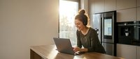 Young woman working on laptop in modern kitchen with natural sunlight streaming through window creating warm atmosphere for remote work and productivity.