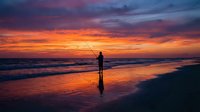 Silhouette of fisherman casting line at sunset on ocean beach with dramatic orange and purple sky reflecting on wet sand and gentle waves rolling ashore.