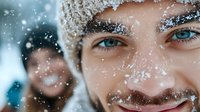 Close up portrait of young Caucasian man with blue eyes and snow-covered face smiling outdoors during winter snowfall, with blurred female figure in background.