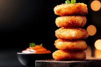 Stack of golden crispy breaded fish cakes garnished with fresh herbs and sauce on dark wooden board against black background with bokeh lights.