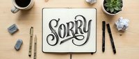 Hand-lettered sorry text in open notebook on wooden desk with coffee, pens, eraser, crumpled paper, and succulent plant for apology concept.