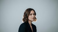 Professional young woman with wavy brown hair in black blazer looking confidently at camera against neutral background for business portraits.