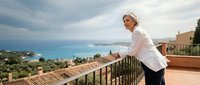 Professional female chef in white uniform standing on Mediterranean terrace overlooking azure sea and coastal landscape for culinary tourism concepts.