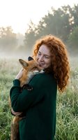 Young redhead woman holding baby fox in green sweater outdoors at sunset with warm golden light and natural meadow background for wildlife conservation content.