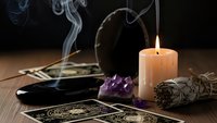 Mystical tarot cards with burning candle, amethyst crystal and sage bundle creating atmospheric smoke on wooden table for spiritual readings.