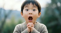 Young Asian boy with surprised expression and open mouth wearing beige sweater outdoors. Child showing amazement and wonder with hands clasped together.