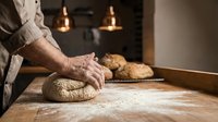Professional baker kneading fresh bread dough on wooden counter in artisan bakery kitchen with warm lighting and freshly baked loaves in background.