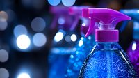 Cleaning spray bottle with blue liquid against blurred bokeh background, bright neon pink sprayer head creates modern aesthetic for cleaning product photography.