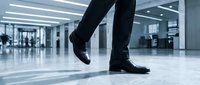 Businessman in black leather shoes walking across modern polished floor in contemporary office lobby with glass partitions and bright lighting.