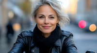 Confident mature woman with silver gray hair smiling outdoors wearing black leather jacket and scarf. Natural lifestyle portrait with soft bokeh background and warm evening light.