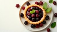 Fresh blackberries and raspberries in golden butter tart with green mint leaves on white ceramic plate, top view composition against light background.