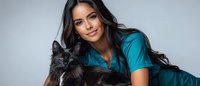 Young Hispanic female veterinarian in teal medical scrubs holding black long-haired cat while smiling at camera against light gray studio background.
