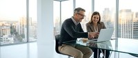 Smiling middle-aged Caucasian businessman and young businesswoman collaborating on laptop in modern office with city view.