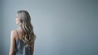 Woman with long wavy silver hair wearing sleeveless top, viewed from behind against neutral blue-gray background, suitable for beauty, hair care, and wellness marketing materials.