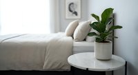 Modern minimalist bedroom interior with green plant on white round nightstand table creating peaceful home decor atmosphere for relaxation and wellness.