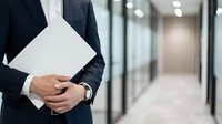 Businessman in dark suit holding white folder with documents in modern office corridor with glass partitions and blurred background.