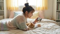 Young Asian woman relaxing on bed with orange tabby cat using smartphone in cozy bedroom with warm morning sunlight streaming through window curtains.