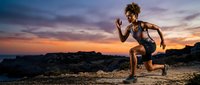 Athletic woman with curly hair in sports attire performing a sprinting motion on rocky terrain during golden hour sunset, suitable for fitness and wellness marketing.