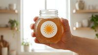 Hands holding glass jar of golden honey with sun logo label in bright modern kitchen for natural organic food packaging and branding concepts.